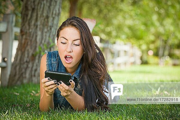 Schockierte gemischtrassige junge Frau textet auf ihrem Handy draußen im Gras liegend