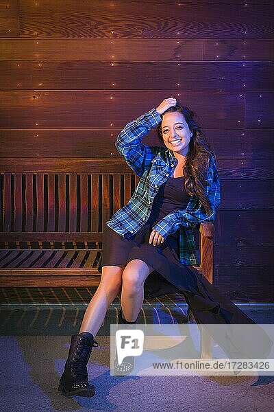 Porträt einer hübschen gemischtrassigen jungen erwachsenen Frau  die auf einer Holzbank vor einem glänzenden Holzwandhintergrund sitzt