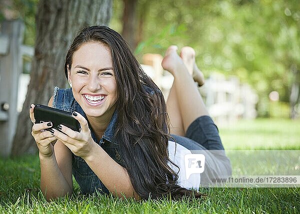 Attraktive glückliche gemischtrassige junge Frau Texting auf ihrem Handy draußen im Gras liegend