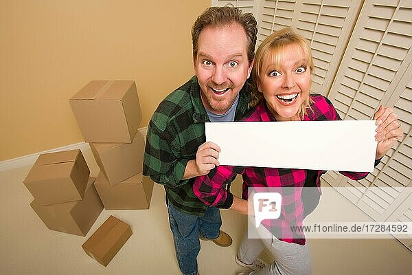 Happy goofy Paar hält leeres Schild im Raum mit gepackten Kisten