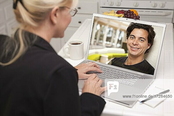 Happy young woman in kitchen using laptop online dating search with portrait of man on screen
