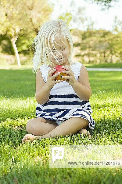 Nettes kleines Mädchen sitzt und ißt Apfel draußen auf dem Gras