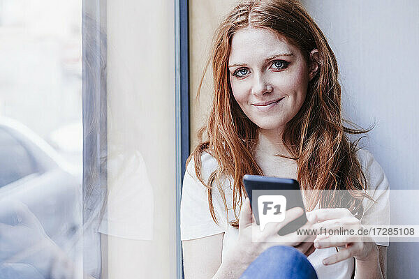 Lächelnde Frau  die ihr Handy hält  während sie zu Hause am Fenster sitzt