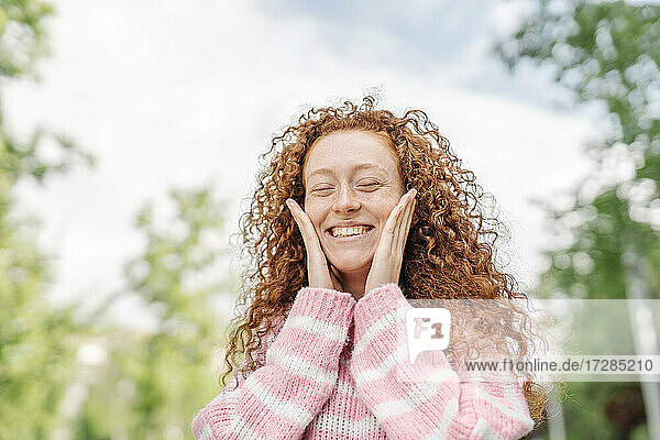Fröhliche rothaarige Frau mit geschlossenen Augen  die ihre Wangen berührt  während sie im Park steht