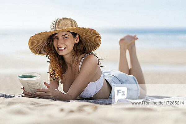 Lächelnde schöne Frau mit Buch liegt auf Handtuch am Strand während sonnigen Tag