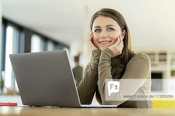 Lächelnde Geschäftsfrau mit Hand am Kinn  die wegschaut  während sie im Büro vor einem Laptop sitzt