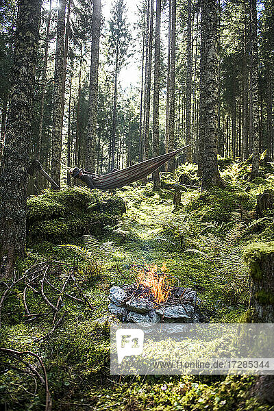 Stone campfire burning in forest with man lying in hammock in background