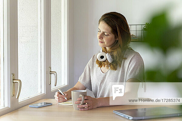 Frau hält Tasse  während sie in einem Buch am Schreibtisch im Heimbüro schreibt
