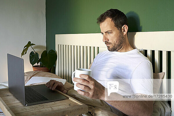 Mann mit Kaffeetasse und Laptop  während er sich zu Hause ausruht