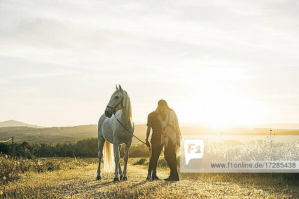Junges Paar  das sich umarmt  während es bei Sonnenuntergang neben einem Pferd auf einem Feld steht