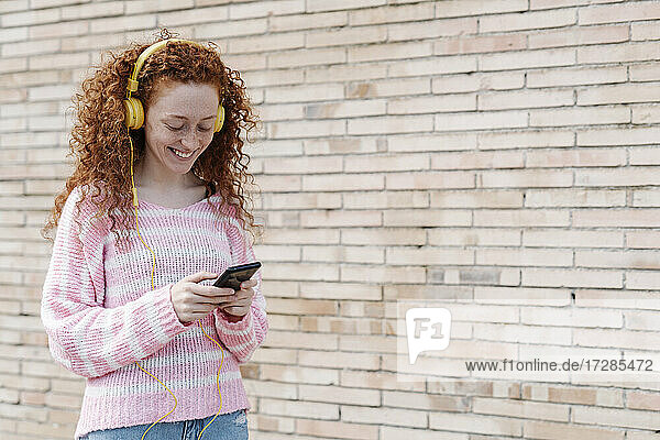 Lächelnde junge Frau  die ein Smartphone benutzt  während sie über Kopfhörer an der Wand Musik hört