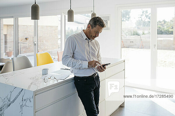 Männlicher Freiberufler arbeitet am Handy  während er zu Hause vor der Kücheninsel steht