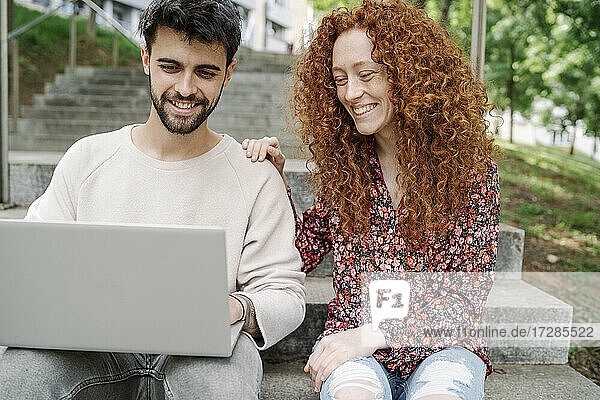 Lächelndes junges Paar  das einen Laptop benutzt  während es auf den Stufen eines öffentlichen Parks sitzt