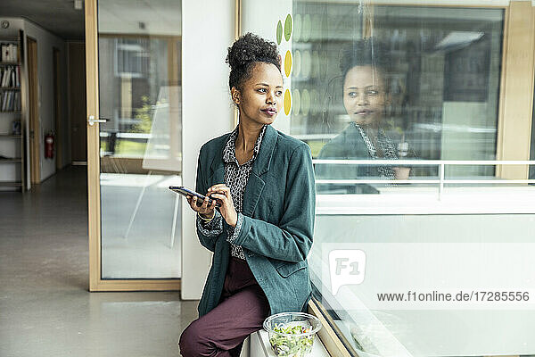 Unternehmerin mit Mobiltelefon  die auf der Fensterbank im Büro sitzt und wegschaut