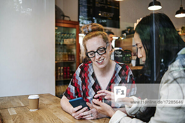 Woman showing smart phone to smiling female friends while sitting at table in cafe