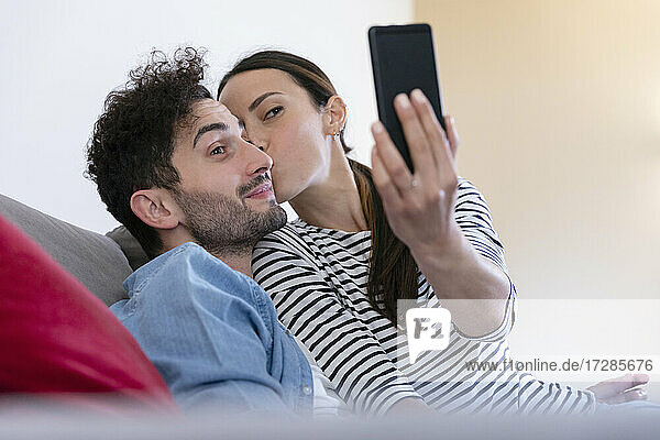 Girlfriend taking selfie through smart phone while kissing on boyfriends cheek at home
