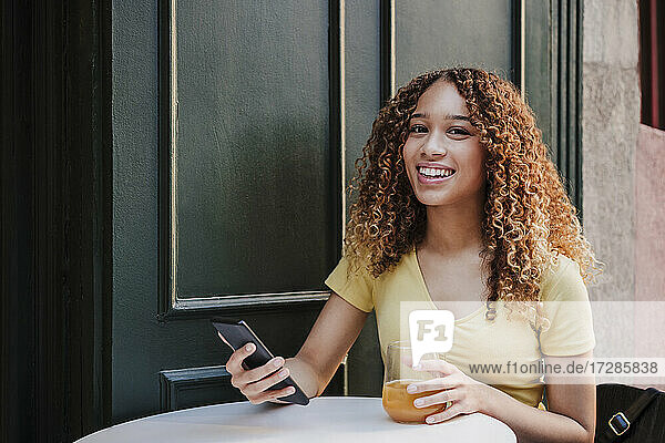 Lächelnde Frau mit Orangensaft  die ihr Smartphone hält  während sie in einem Straßencafé sitzt
