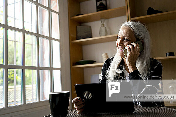 Lächelnde Frau mit digitalem Tablet  die zu Hause mit ihrem Mobiltelefon spricht