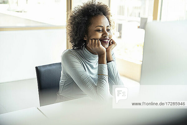Lächelnde weibliche Fachkraft mit Hand am Kinn  die auf einen Computer im Büro schaut