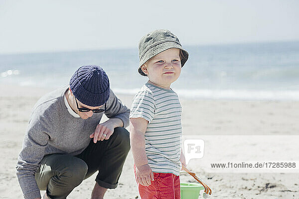 Niedlicher kleiner Junge  der wegschaut  während er neben seinem Vater am Strand steht