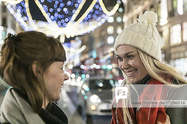 Smiling blond woman wearing knit hat looking at female friend while talking on Christmas