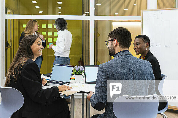Smiling male and female entrepreneurs with laptop discussing during meeting with coworkers in background at office