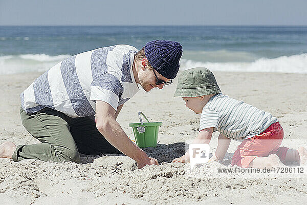 Lächelnder Mann gräbt mit seinem Sohn am Strand bei Sonnenschein im Sand