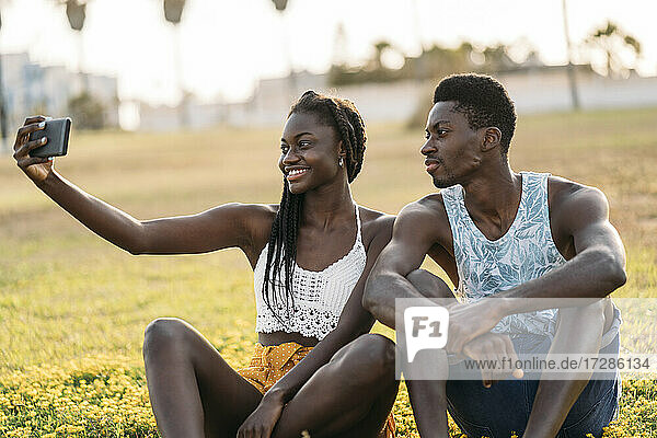 Paar nimmt Selfie durch Handy beim Sitzen auf Wiese während Sonnenuntergang