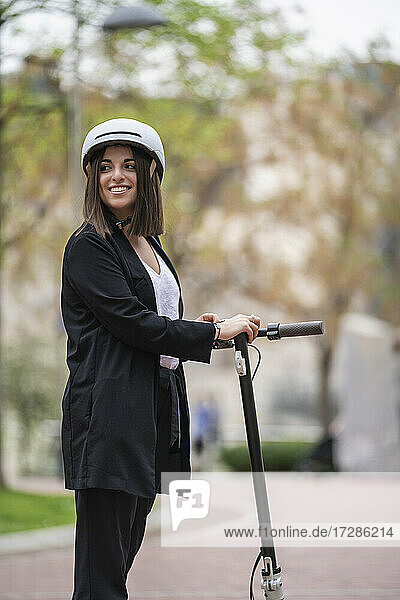 Smiling young businesswoman with electric push scooter looking away while standing on footpath