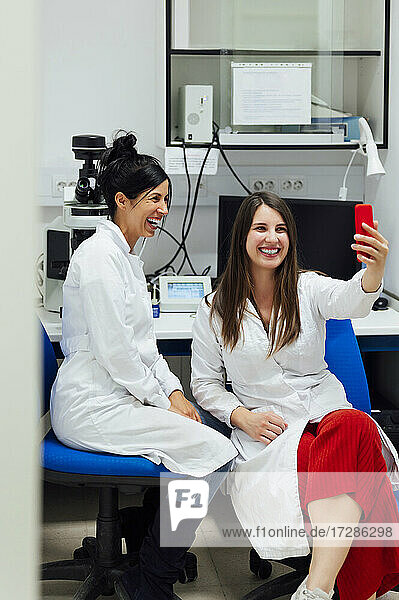 Fröhliche junge Medizinerin  die ein Selfie mit einem Kollegen im Labor über ihr Smartphone macht