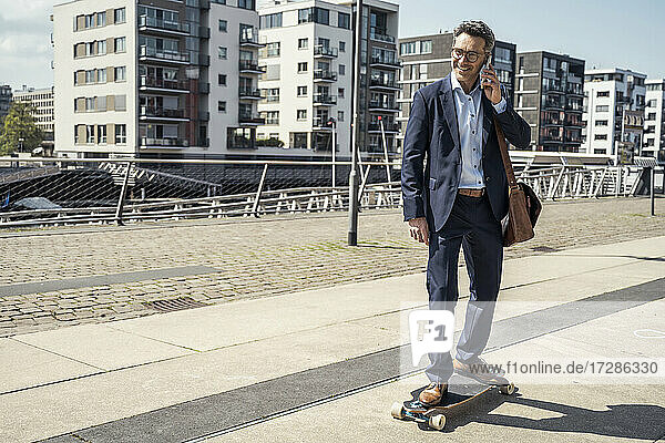 Lächelnder männlicher Unternehmer  der mit seinem Handy telefoniert  während er in der Stadt Skateboard fährt