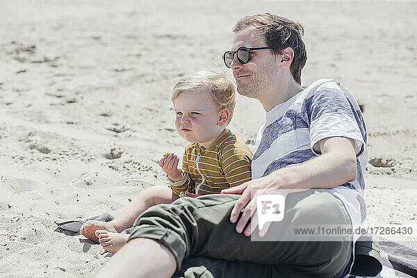 Smiling man leaning by son sitting at beach during sunny day