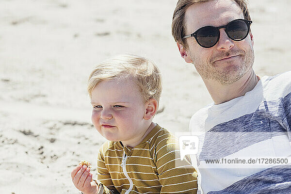 Lächelnder Mann mit Sonnenbrille sitzt mit seinem Sohn am Strand