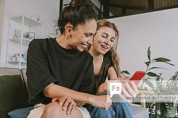 Lächelnde Freundinnen  die auf ihr Smartphone schauen  während sie zu Hause sitzen