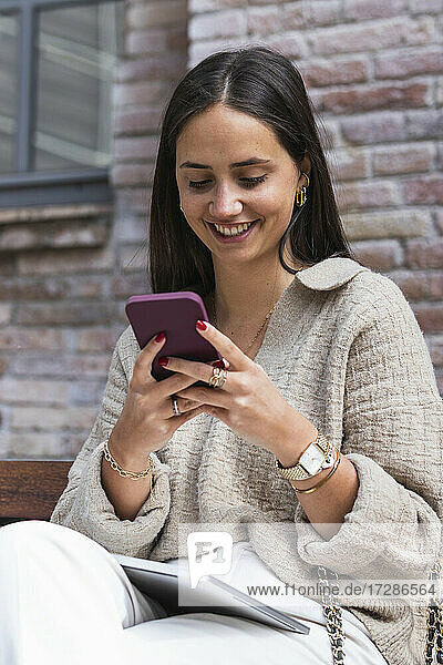 Lächelnde schöne Frau  die auf einer Bank ein Mobiltelefon benutzt