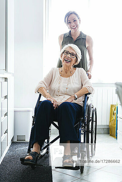 Fröhliche  behinderte Seniorin im Rollstuhl sitzend  neben ihrer Enkelin in der heimischen Küche stehend