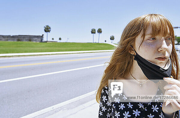Rothaarige junge Frau mit Gesichtsschutzmaske bei COVID-19  die an der Straße steht und wegschaut