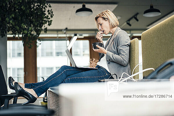 Unternehmerin beim Essen  während sie im Büro einen Laptop benutzt