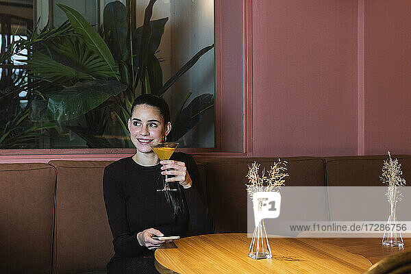 Smiling woman with mobile phone and martini glass sitting at restaurant