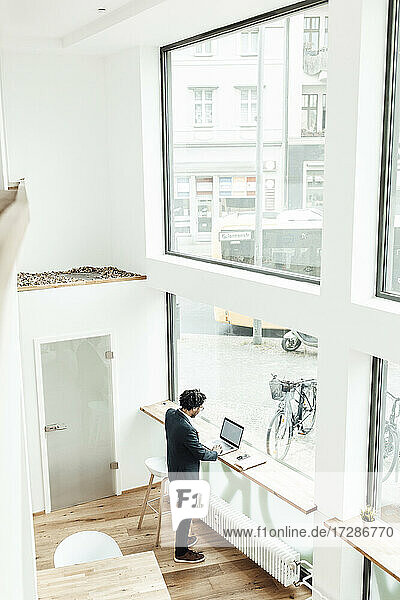 Männlicher Unternehmer  der einen Laptop benutzt  während er vor einem Glasfenster im Büro steht