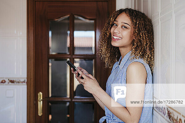 Lächelnde Frau mit Mobiltelefon  die sich zu Hause an eine gekachelte Wand lehnt