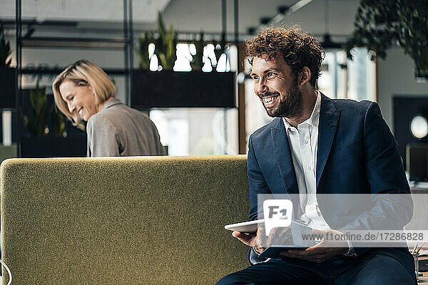 Lächelnder Geschäftsmann  der ein digitales Tablet hält  während er mit einem Kollegen im Hintergrund im Büro sitzt