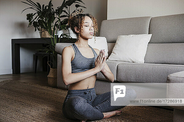 Junge Frau mit verschränkten Händen beim Meditieren auf einem Teppich zu Hause sitzend