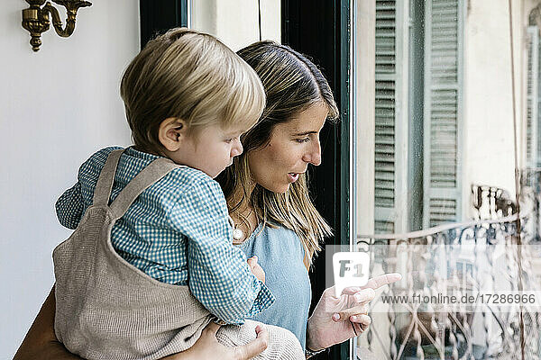 Frau trägt ihren Sohn und schaut durch das Fenster ihres Hauses