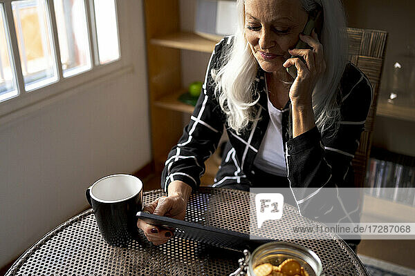 Frau mit digitalem Tablet  die am Tisch im Wohnzimmer sitzt und mit ihrem Mobiltelefon spricht