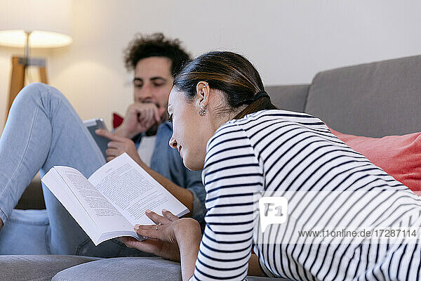 Eine Frau liest ein Buch und ein Mann benutzt ein digitales Tablet auf dem Sofa zu Hause