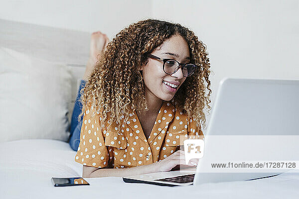 Junge Frau mit Brille benutzt einen Laptop  während sie zu Hause auf dem Bett liegt