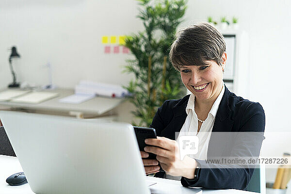 Fröhliche weibliche professionelle Textnachrichten über ein Smartphone  während sie am Schreibtisch im Büro sitzt