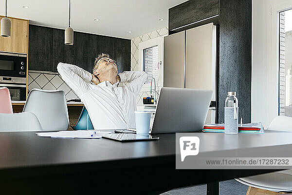Reifer Geschäftsmann mit Händen hinter dem Kopf am Tisch sitzend im Heimbüro