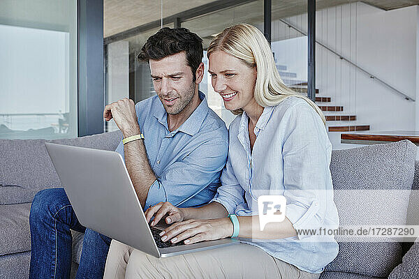 Lächelnde blonde Frau  die einen Laptop benutzt  während ein Mann auf dem Sofa im Wohnzimmer sitzt